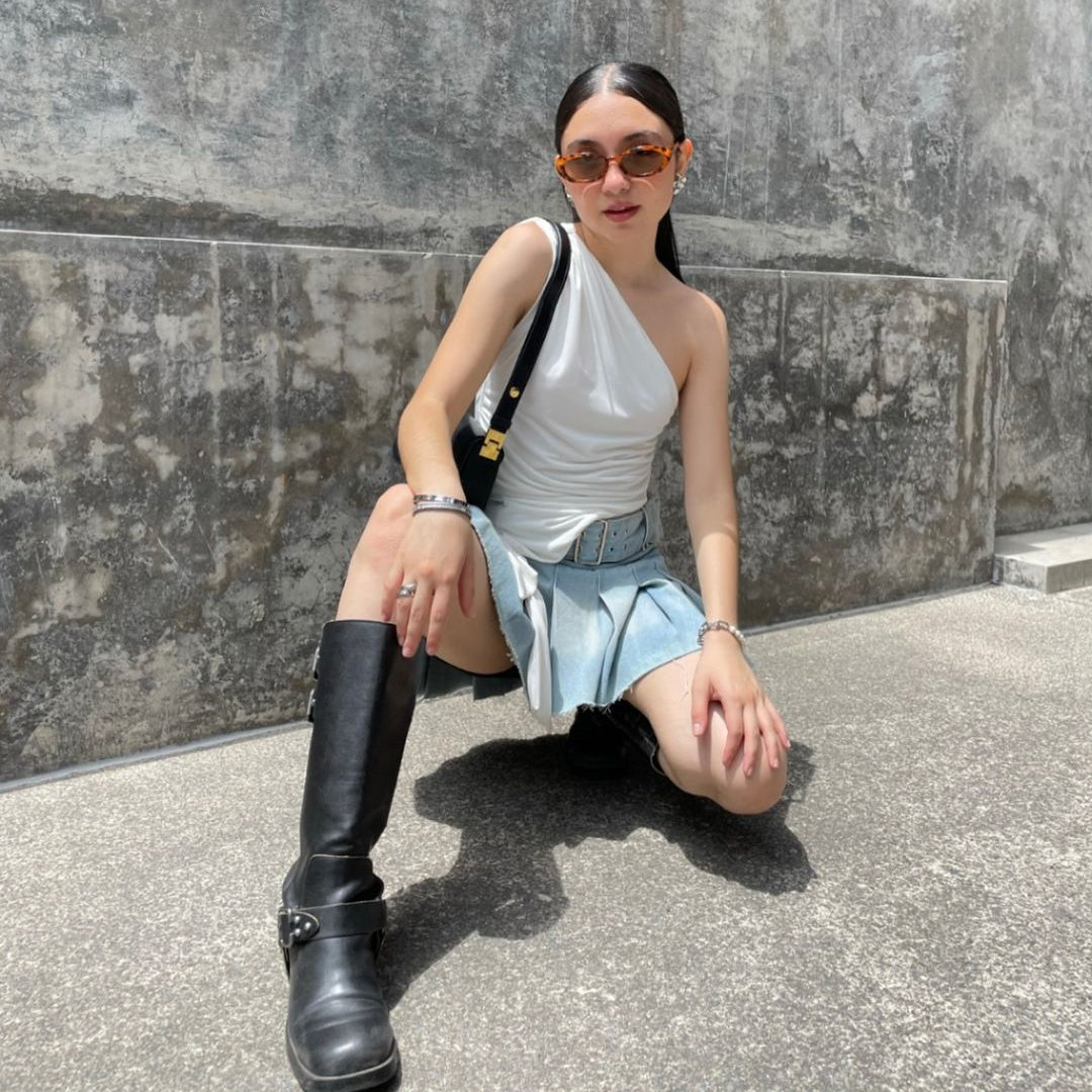 Person posing in a white top, denim skirt, and black boots against a concrete wall.